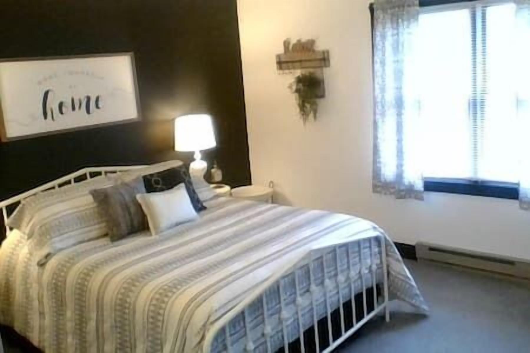 Une chambre à coucher comprenant un lit, une table de chevet, une lampe, une fenêtre avec des rideaux et une œuvre d’art accrochée au mur.