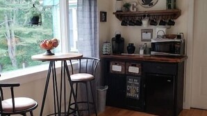 Une cuisine chaleureuse avec un plan de travail en bois, une machine à café et un réfrigérateur. On y trouve deux tabourets et une table agrémentée d’un vase de fleurs.