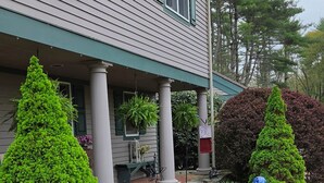 Une maison avec un porche, des fenêtres bordées de vert et un jardin bien entretenu, agrémenté de diverses plantes et d’un abreuvoir pour les oiseaux.