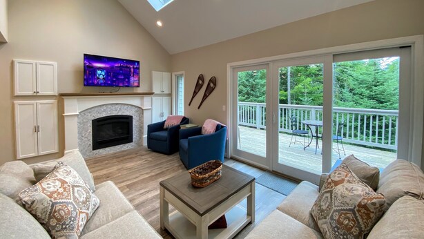 A living room with a fireplace, sofas, a television, and a balcony with a view of trees.