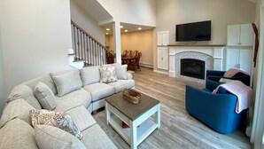 A spacious living room with a sectional sofa, a blue armchair, a fireplace, a TV mounted above it, and a staircase leading to an upper level.