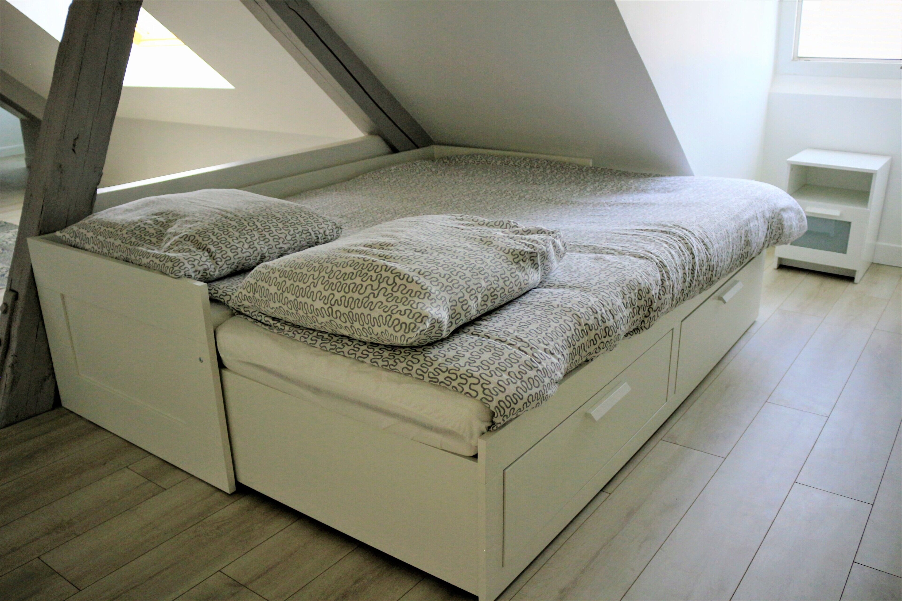 Un dormitorio estilo loft con techo inclinado, una cama con ropa de cama estampada y una mesita de noche pequeña y blanca.