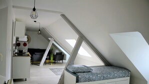 A modern loft bedroom with a sloped ceiling, a bed with patterned bedding, a white dresser, and a dining area in the background. - Apartment center Rouen (Rouen)