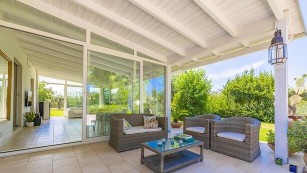 Un patio couvert aménagé avec des meubles en osier, une table basse en verre et des plantes en pot.