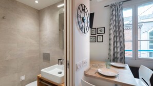 A modern bathroom with a toilet, sink, and shower. There is a dining area with a table, chairs, and a clock on the wall. - Le Petit Botté - Studio plein cœur de Rennes (Rennes)