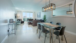 Un salon moderne avec une table à manger en verre, des chaises noires et un canapé. Un grand miroir orne le mur et un luminaire suspendu se trouve au-dessus de la table.