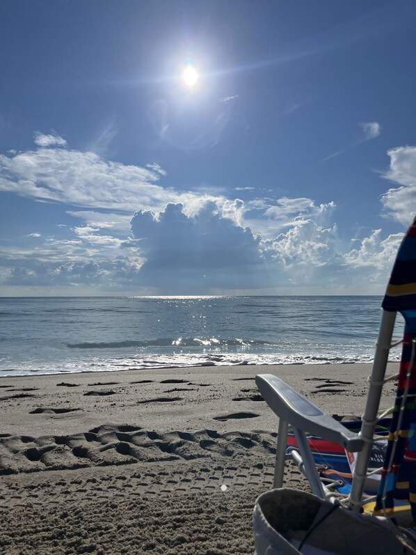 A beach with a clear sky, a sun shining, and a chair and bucket on the sand.