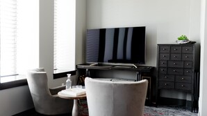 A hotel room with a flat-screen TV mounted on the wall, a small table with a bottle, two chairs, and a dark wooden cabinet with drawers.