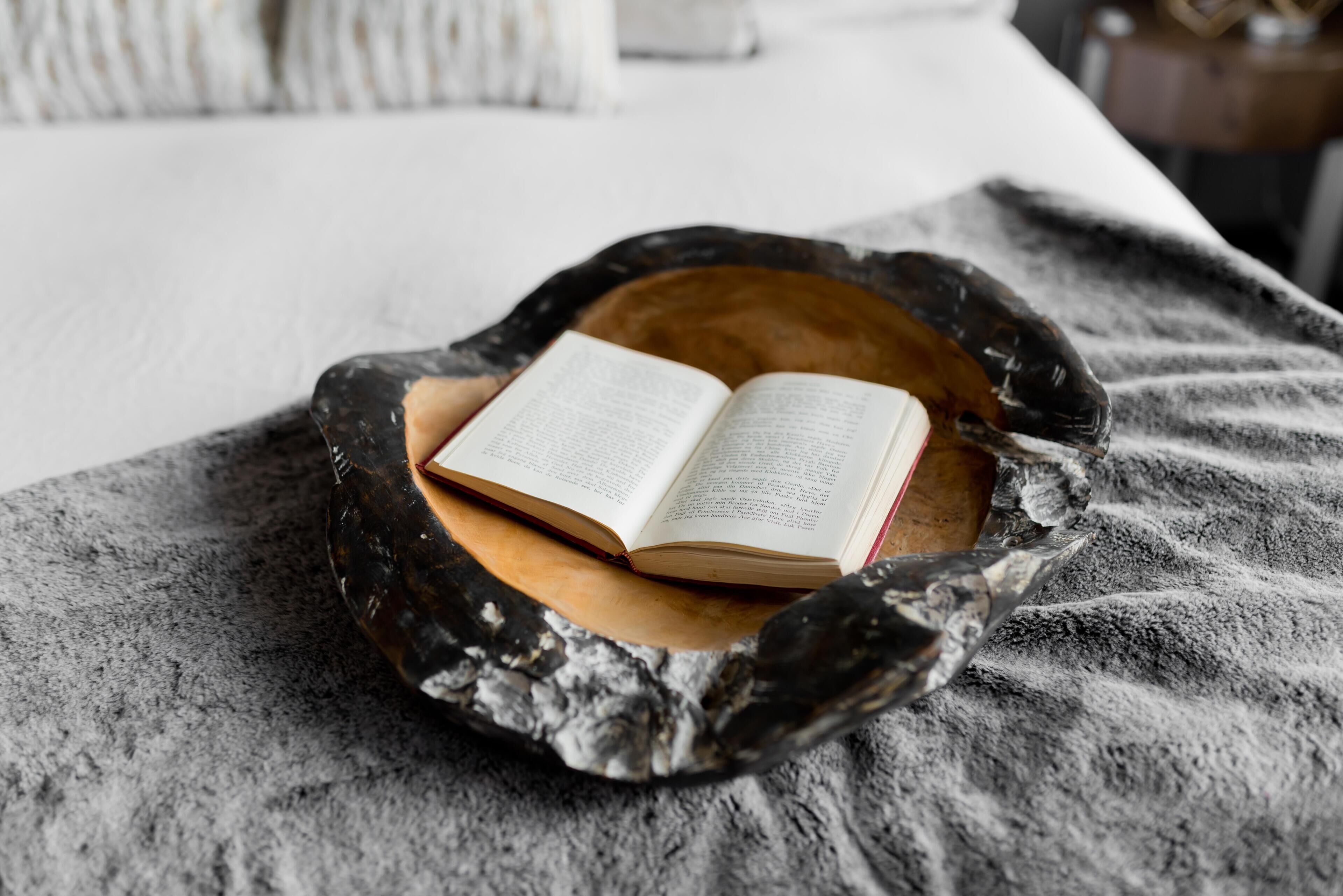 A book resting on a wooden bowl placed on a bed with a gray blanket.