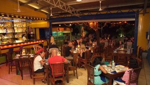 A restaurant with wooden tables and chairs, a bar area with shelves of bottles, and a view of the outdoor seating area with string lights. - New York Guest House (Negombo)