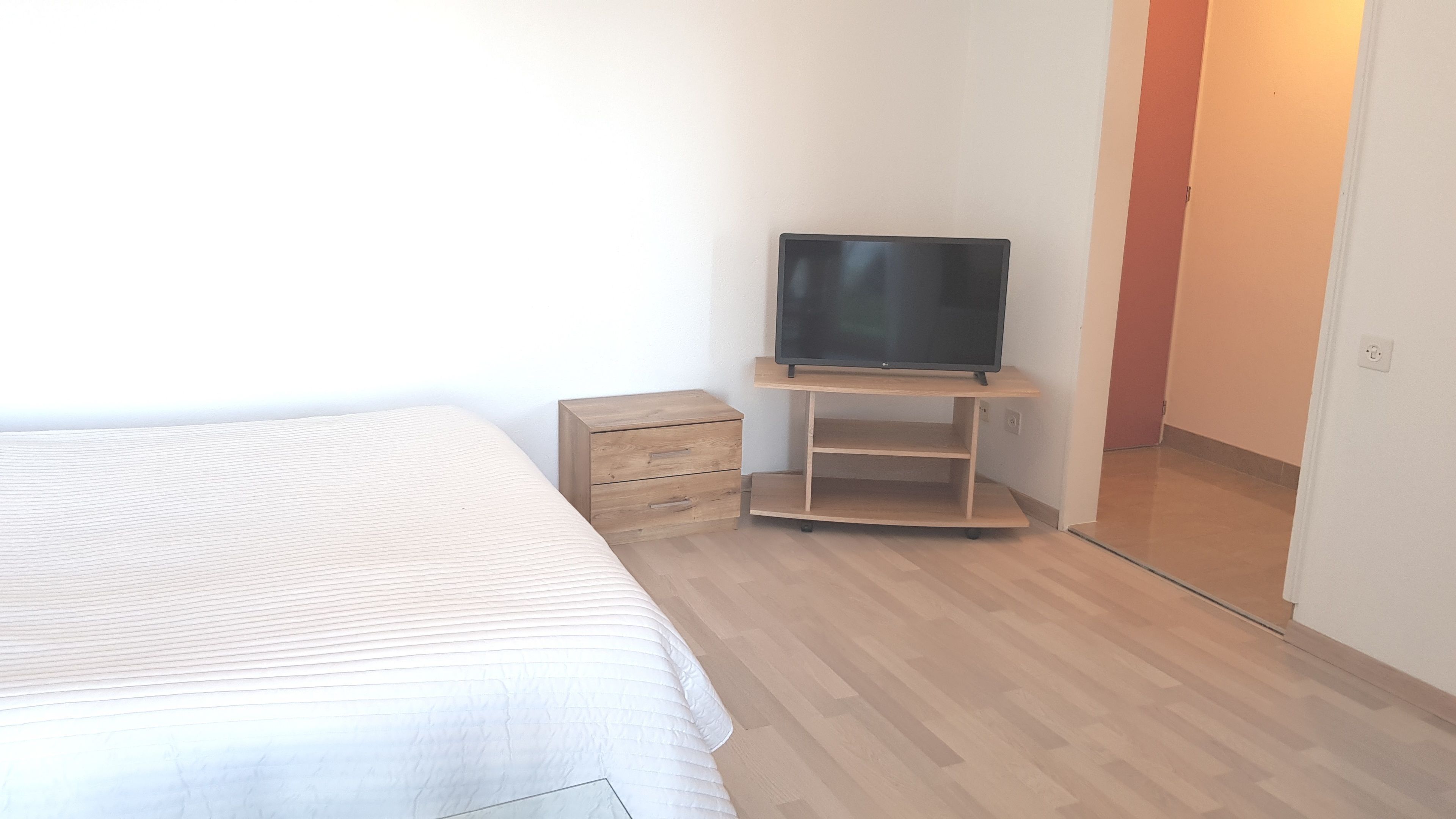 Un dormitorio con una cama, un televisor sobre un mueble bajo, un pequeño armario de madera y una puerta que da a otra habitación.