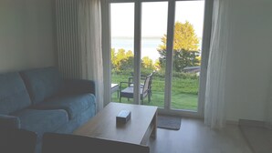 A room with a sofa, a wooden table, and a view of a garden and water through large windows. - Littoral Lac (La Grande-Béroche)