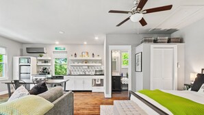 A modern, well-lit room with a kitchenette, a dining area, and a bedroom with a large bed and a ceiling fan.