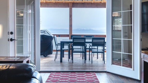 An open door leading to a deck with a table and chairs, overlooking a body of water and mountains. - Incredible View, Privacy & Private hot tub! (Homer)