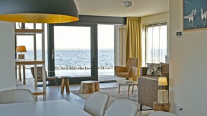 Un salon moderne doté d’une grande fenêtre donnant sur la mer, un coin repas avec une table blanche et un espace salon confortable.