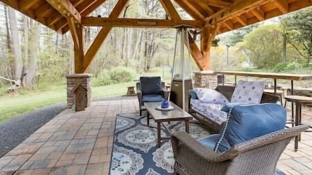 A covered patio with a wooden roof, stone pillars, a patterned rug, and outdoor furniture including a sofa and chairs.