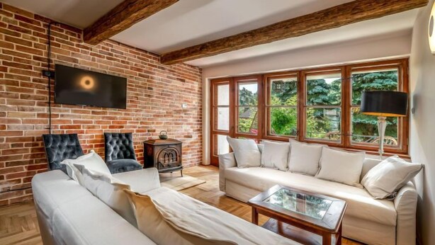 A living room with a brick wall, wooden beams, a large window with a view of greenery, a sofa, a coffee table, and a television.