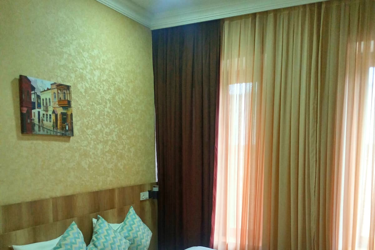 A hotel room with a bed, pillows, a nightstand, a painting on the wall, and curtains.