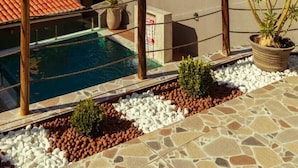 A pool area with a tiled roof, a small building with a cross on top, and a hill with greenery in the background.