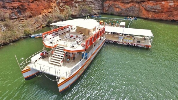 A large riverboat with multiple decks and a covered upper deck, docked at a river with rocky cliffs in the background.