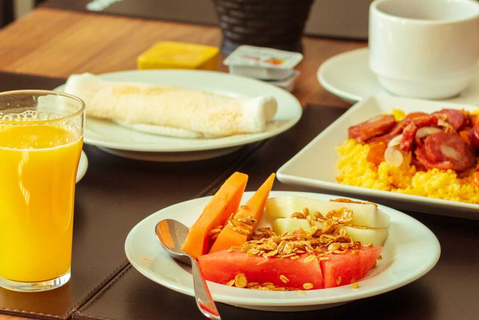 A breakfast setting with a glass of orange juice, a plate with a fruit salad, a plate with scrambled eggs and bacon, and a plate with a rolled omelette.