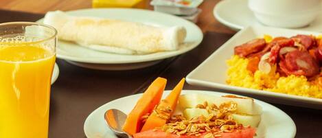 A breakfast setting with a glass of orange juice, a plate with a fruit salad, a plate with scrambled eggs and bacon, and a plate with a rolled omelette.