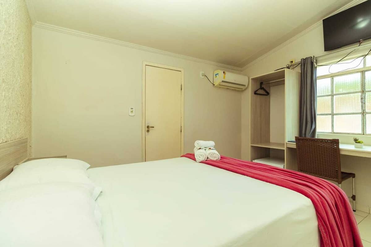 Ein Hotelzimmer mit Bett, Kleiderschrank, Schreibtisch mit Stuhl, Klimaanlage und einem Vorhangfenster.