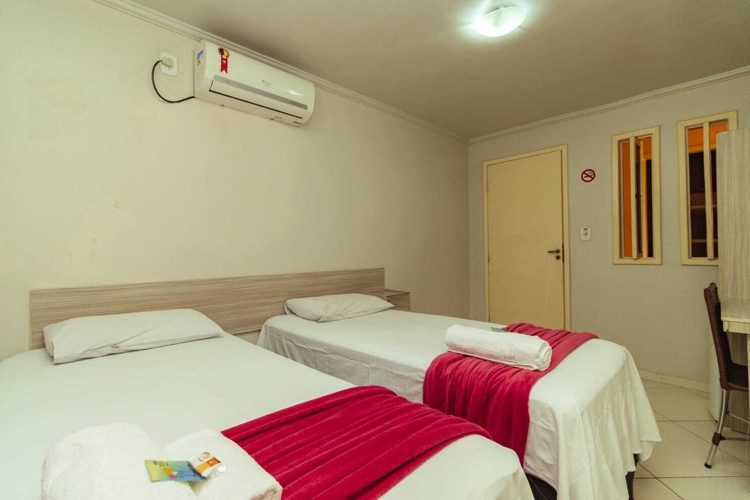 Quarto de hotel com duas camas, uma escrivaninha, uma cadeira, ar condicionado e uma porta com placa de proibido fumar.