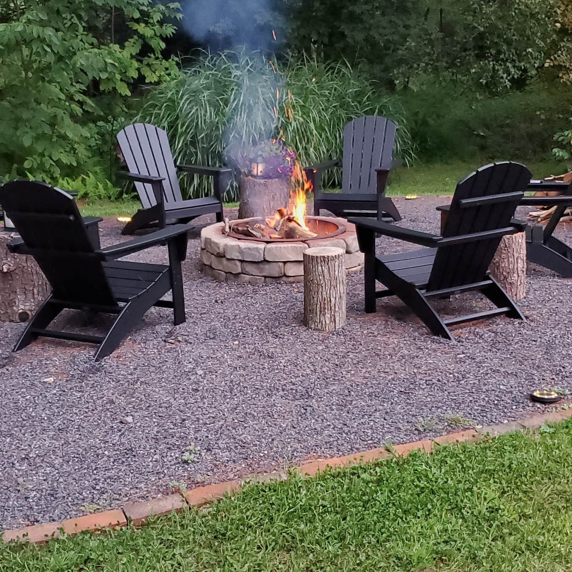 A fire pit with four Adirondack chairs around it, set on a gravel surface with a grassy border.
