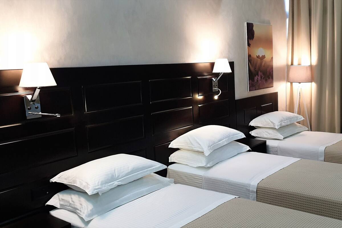 Une chambre d’hôtel avec trois lits simples, une tête de lit sombre, deux lampes fixées au mur, un tableau et une fenêtre avec des rideaux.