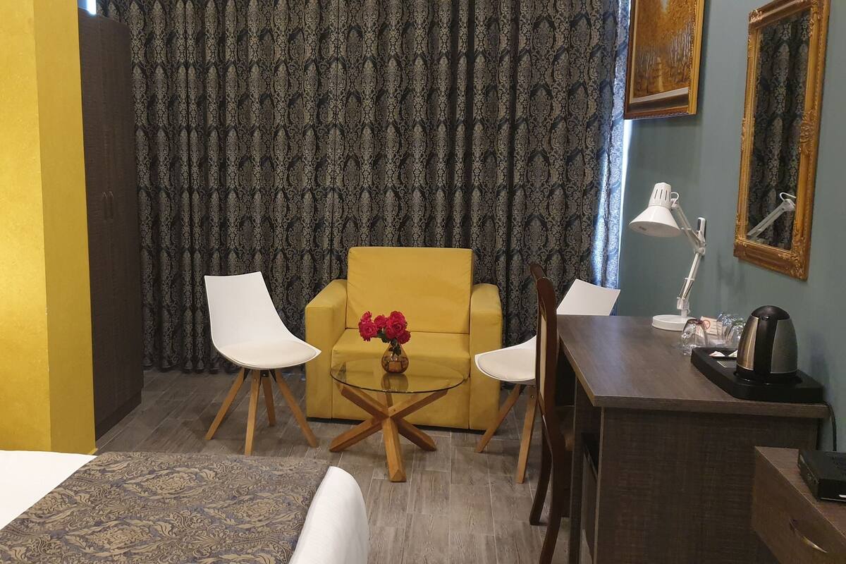 Une chambre d’hôtel avec un lit, un fauteuil jaune, deux chaises blanches, une petite table ronde ornée d’un vase de fleurs, un bureau avec une lampe et un grand miroir.