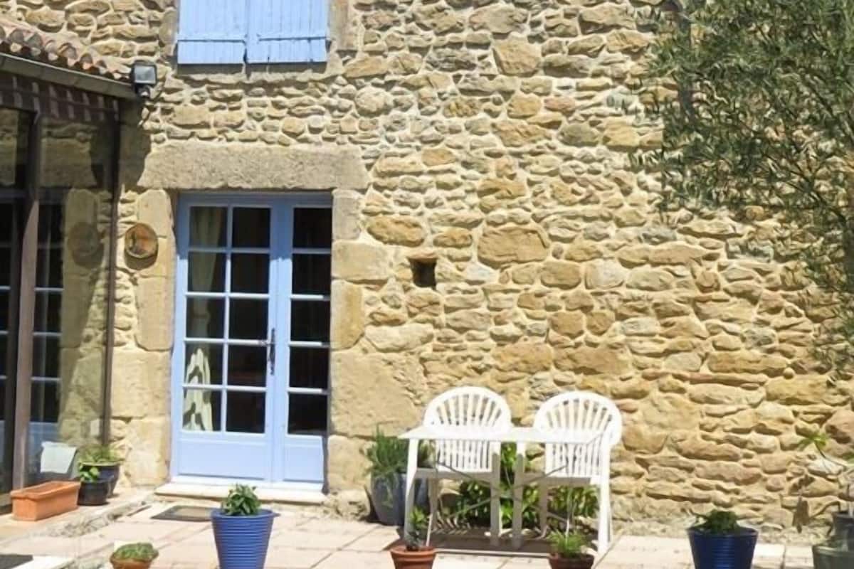 Ein Steinhaus mit blauer Tür und Fensterläden, eine kleine Terrasse mit zwei weißen Stühlen und Topfpflanzen.