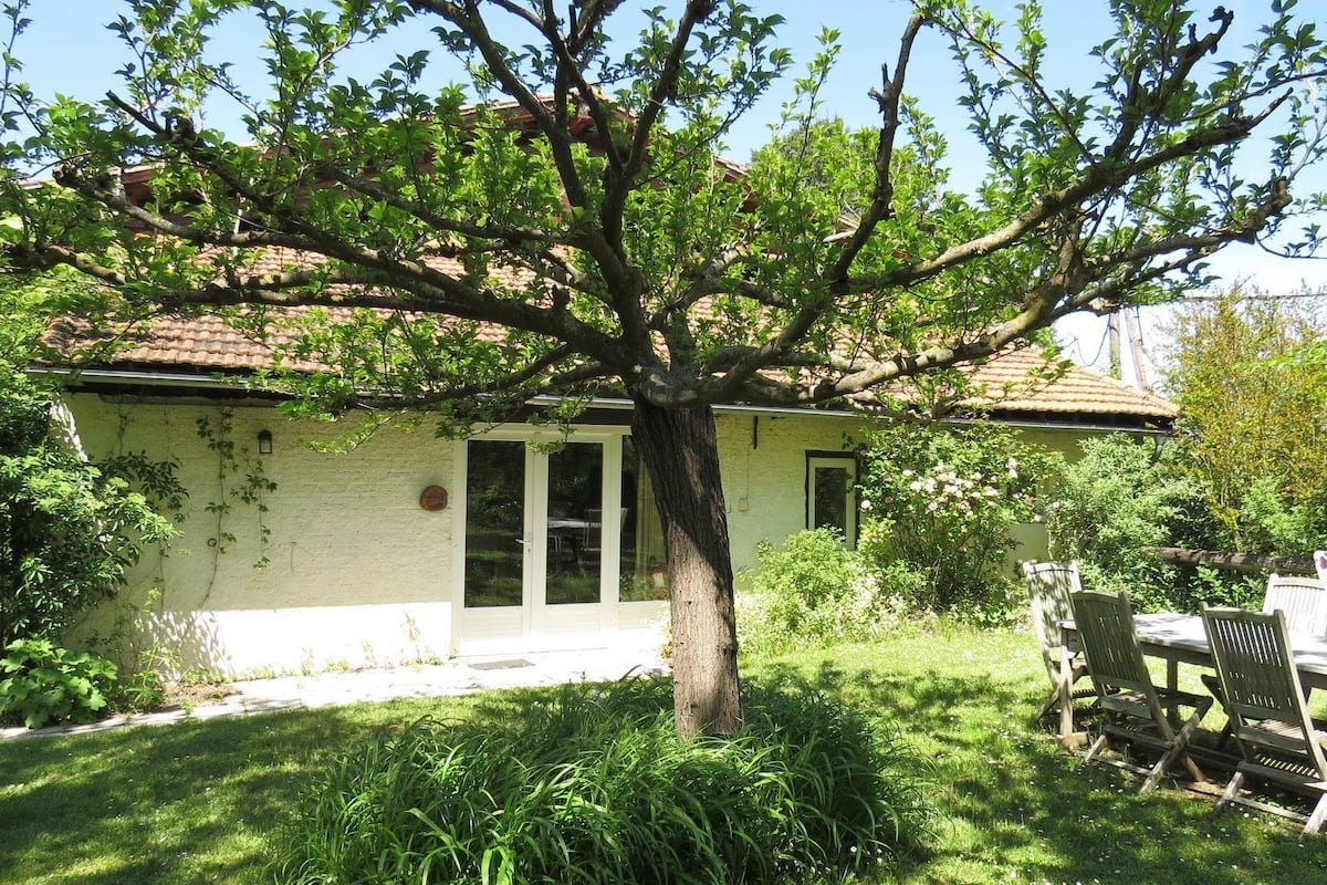Ein kleines Haus mit einem Ziegeldach, einer Haustür und einem Fenster. Vor dem Haus steht ein Baum, und rechts daneben ein Picknicktisch mit Stühlen.