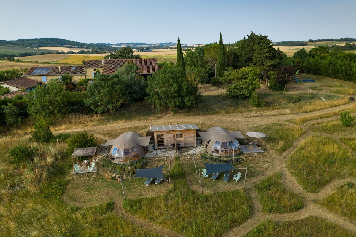 Ländliche Szenerie mit einem kleinen Haus und zwei kuppelförmigen Bauwerken, möglicherweise Jurten, mit Sitzgelegenheiten im Freien und einem Weg zu ihnen.