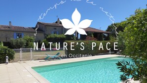 A swimming pool with a clear blue water surface, surrounded by a tiled deck. In the background, there is a stone building with a tiled roof and a fence with a gate. - Nature's Pace Holidays (Montmaur)