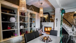 Ein Essbereich mit einem weißen Tisch, schwarzen Stühlen und einer Chandelierleuchtre. Ein Kamin mit sichtbarem Feuer und eine Treppe mit einer Holzhandläufung sind ebenfalls vorhanden.