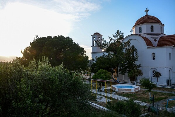 A church with a bell tower and a dome, surrounded by trees and a fenced playground. - Traditional one bedroom village home (Ierapetra)