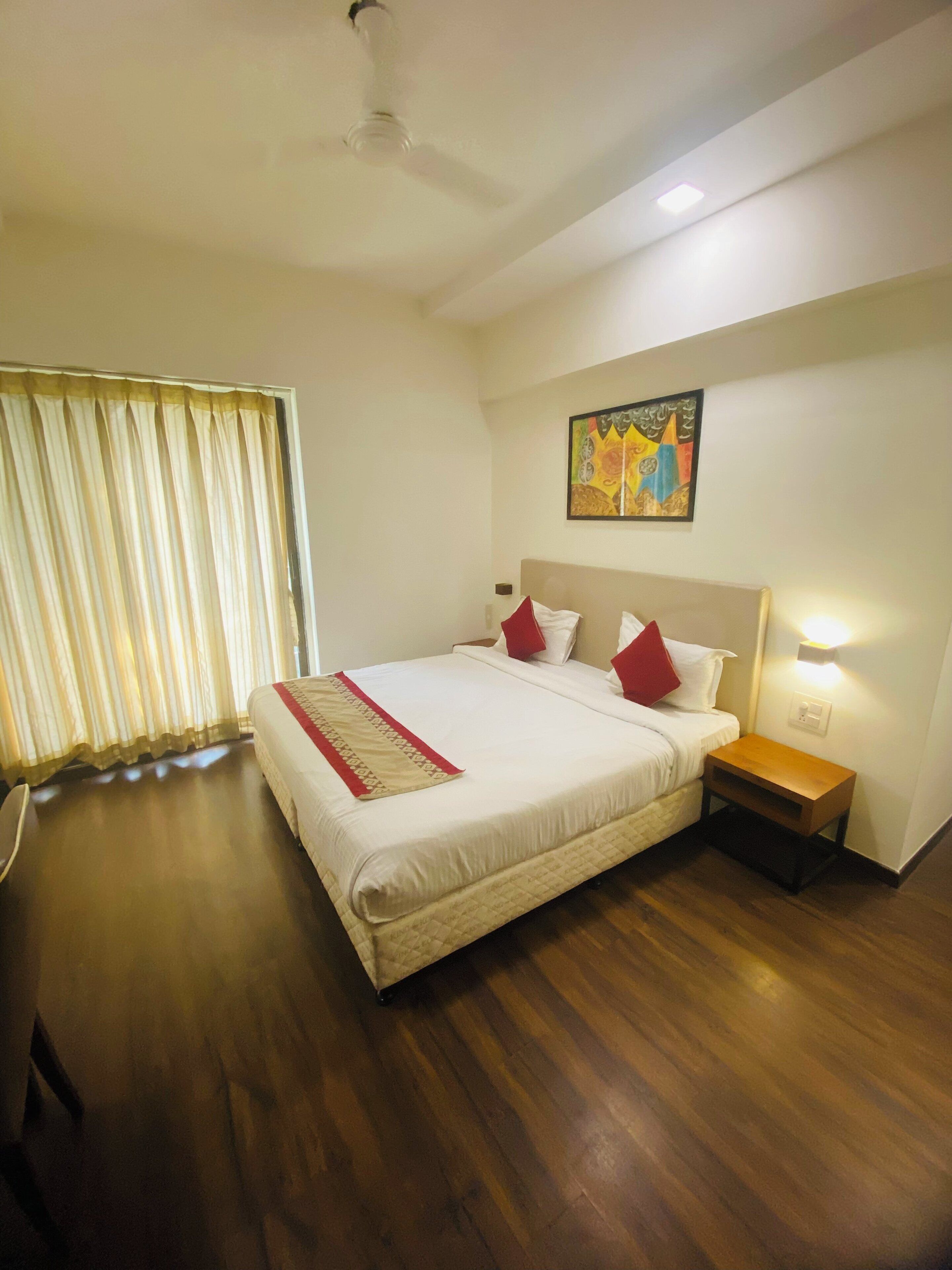 Quarto de hotel com uma cama grande, uma mesinha de cabeceira, um ventilador de teto e uma janela com cortinas.