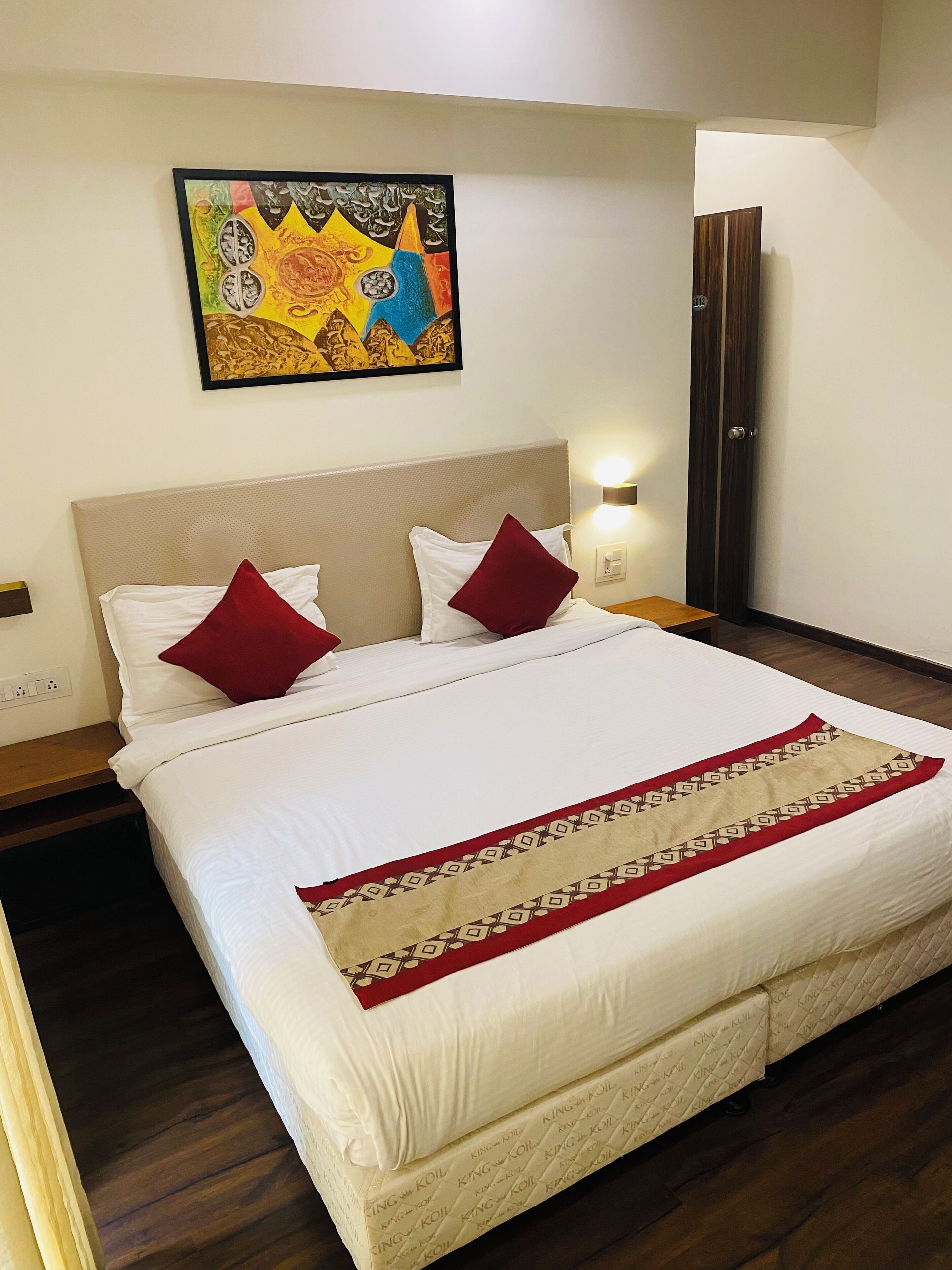 A neatly made bed with white linens and red pillows, a decorative throw, and a painting above it.