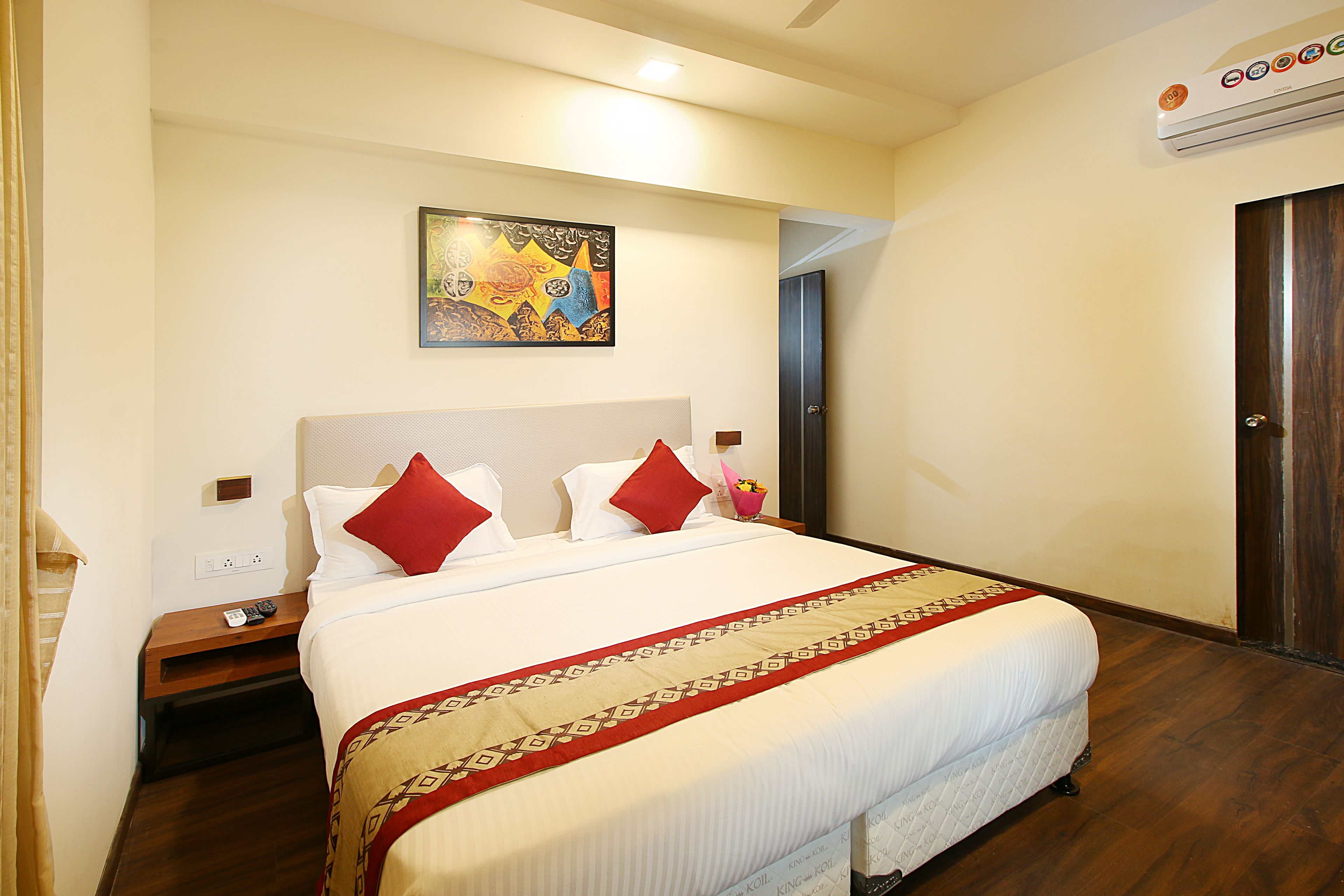 Ein Hotelzimmer mit einem großen Bett, einem Gemälde an der Wand, einem Nachttisch mit Fernbedienung und einer Tür rechts.