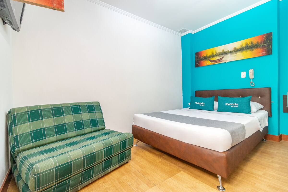Une pièce comprenant un lit, un canapé à carreaux verts, un mur d’accent de couleur turquoise et un tableau représentant un paysage fluvial.