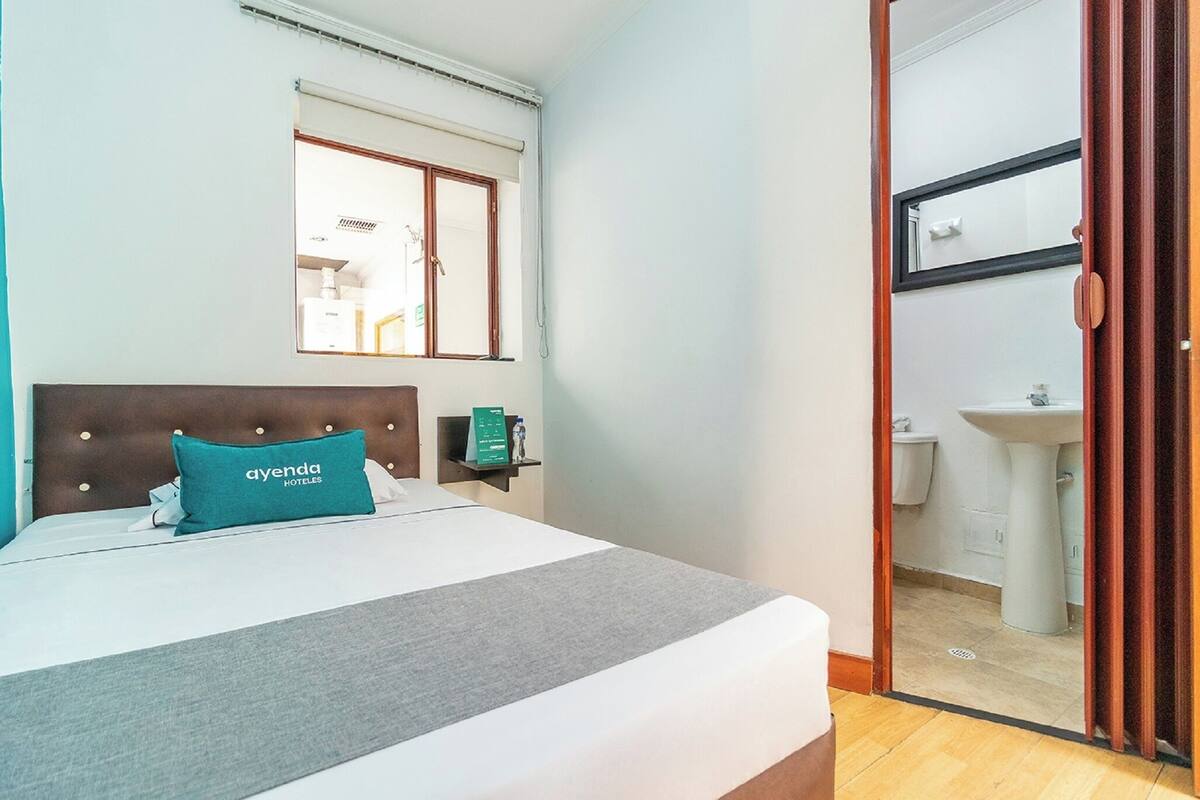Une chambre d’hôtel avec un lit, un oreiller orné du texte « AYENDA HOTELES », une table de chevet avec un panneau vert, une fenêtre avec des stores et une vue sur une salle de bain.
