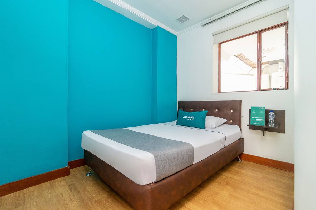 Une chambre d’hôtel avec un lit, des murs de couleur turquoise, un plancher en bois, une fenêtre et un panneau d’information fixé au mur.