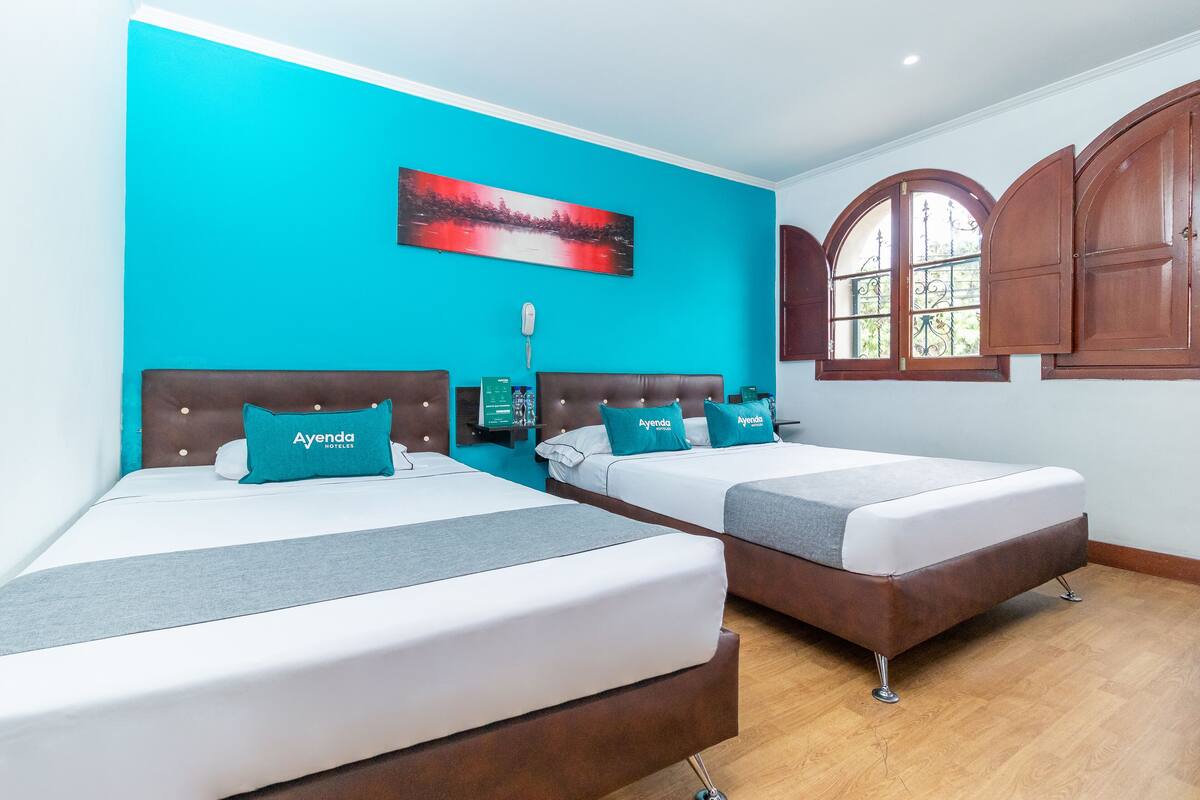 Deux lits avec des têtes de lit marron, du linge de lit blanc et des coussins turquoise, dans une pièce aux murs turquoise et au plancher en bois.