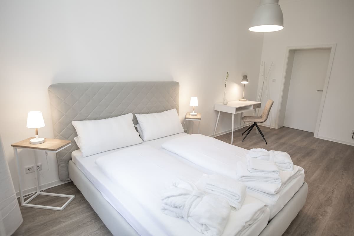 A neatly made bed with white linens and a folded robe, a bedside table with a lamp, a desk with a chair, and a hanging light fixture.