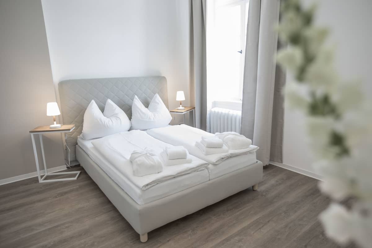A neatly made bed with white linens and pillows, a bedside table with a lamp, and a view of a window with curtains.