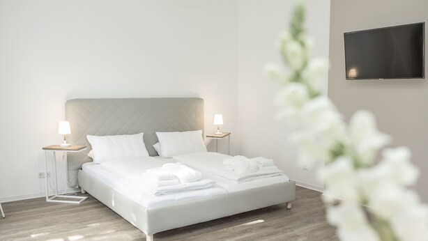 A modern bedroom with a bed, two bedside tables, a flat-screen TV, and a wooden floor.