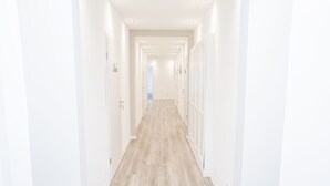 A long, narrow hallway with white walls and wooden flooring, leading to a bright room at the end.
