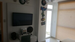 Een kamer met een flatscreen televisie aan de muur, een klein kastje met een computer en een lamp, en een raam met zonwering.