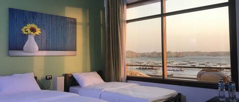 A hotel room with two beds, a painting of sunflowers, a view of a harbor, and a water bottle on a cabinet.